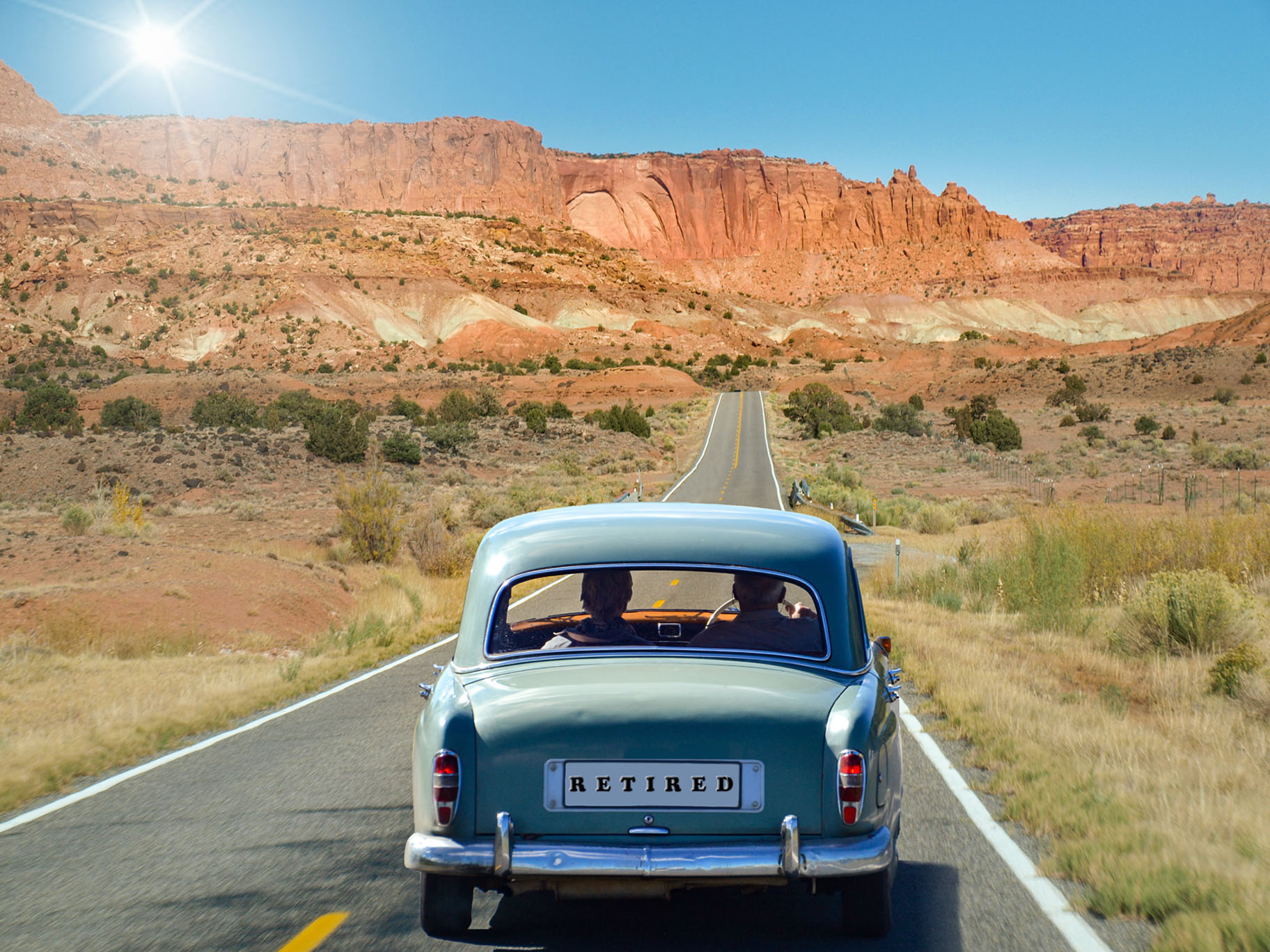 Old man and woman driving in an oldtimer on a long clear road with red mountains in background. The license number says "Retired".