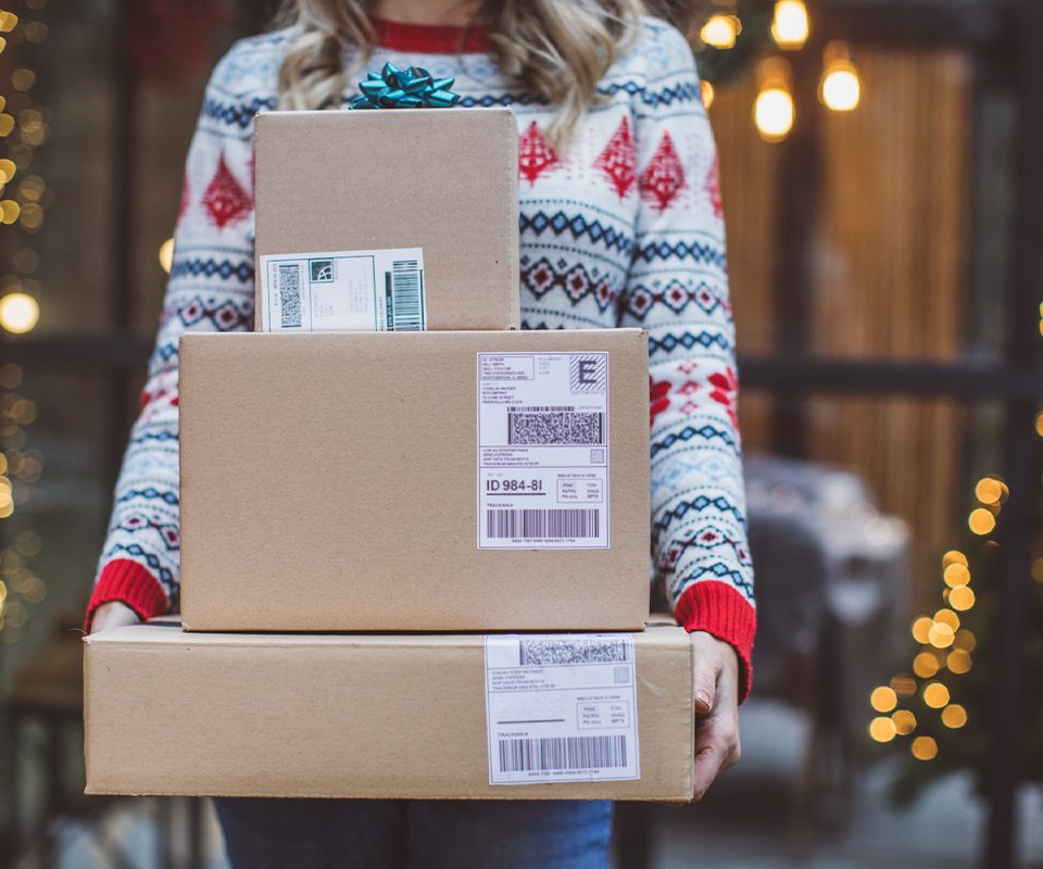 Woman wearing a festive sweater holding a stack of mailed packages with holiday wrapping
