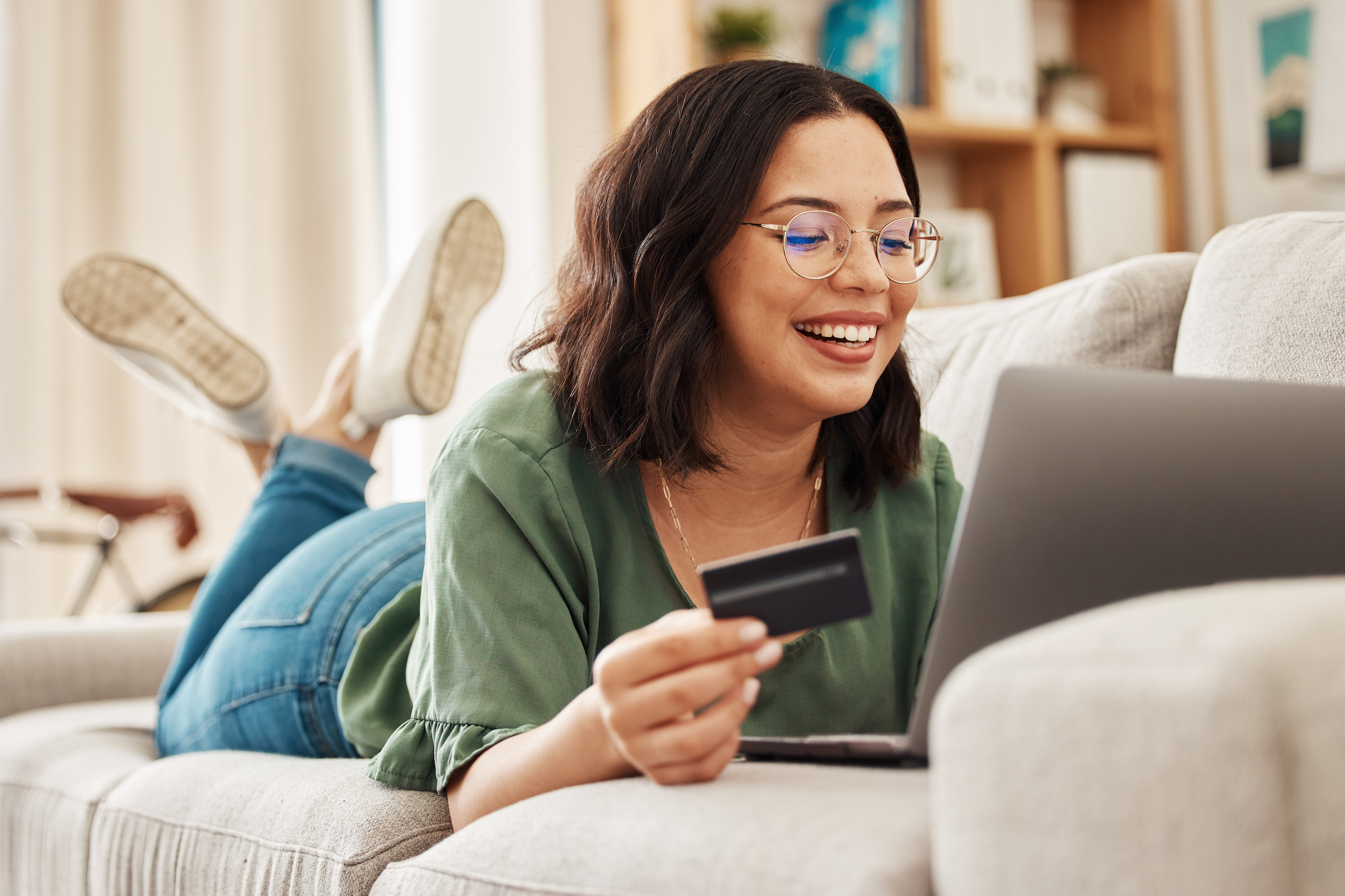 A woman is laying on the couch using her debit card to make purchases on a laptop knowing she's earning debit card rewards.
