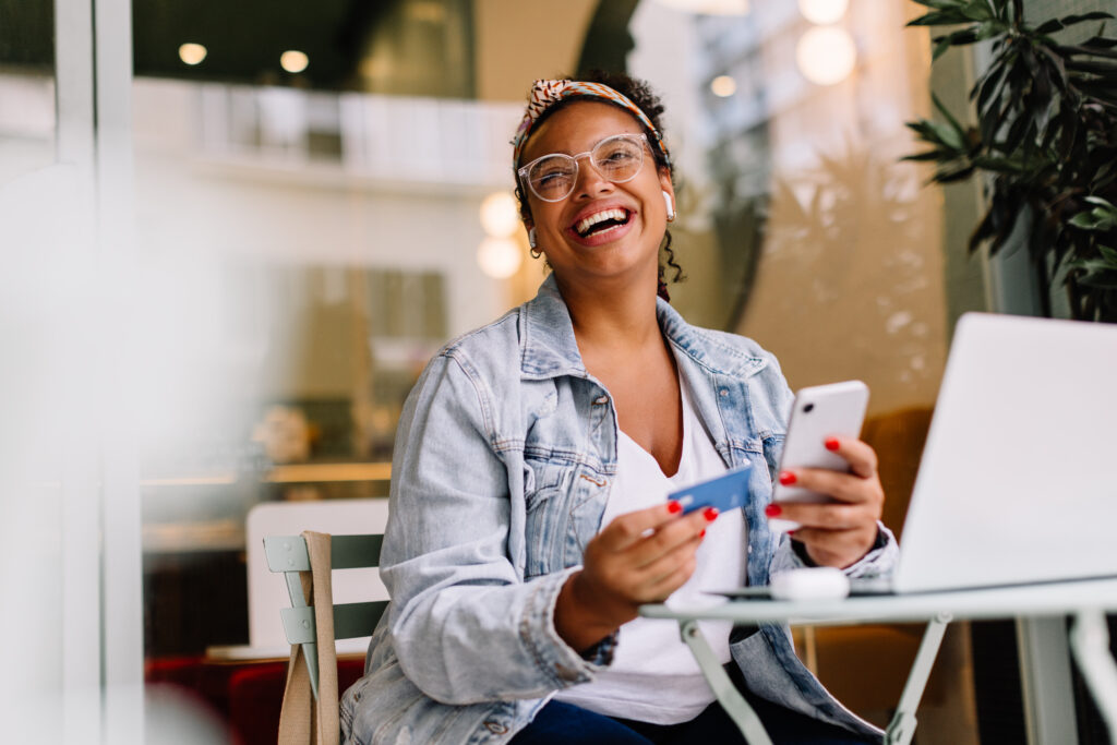 Young woman in a café happily uses her smartphone for online shopping and mobile banking, paying with a credit union credit card. Woman highlighting the convenience of digtal benefits from a credit union.