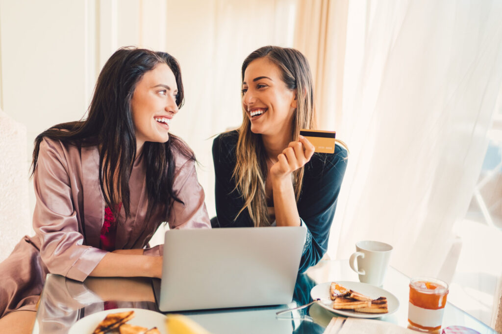 two smiling women sitting side by side at a cafe table with an open laptop, coffee, and breakfast
