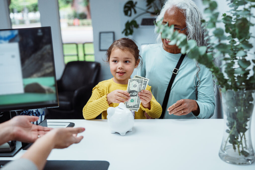 A cheerful five-year-old talks with a female bank teller while depositing money from her piggy bank into a savings account at a credit union.