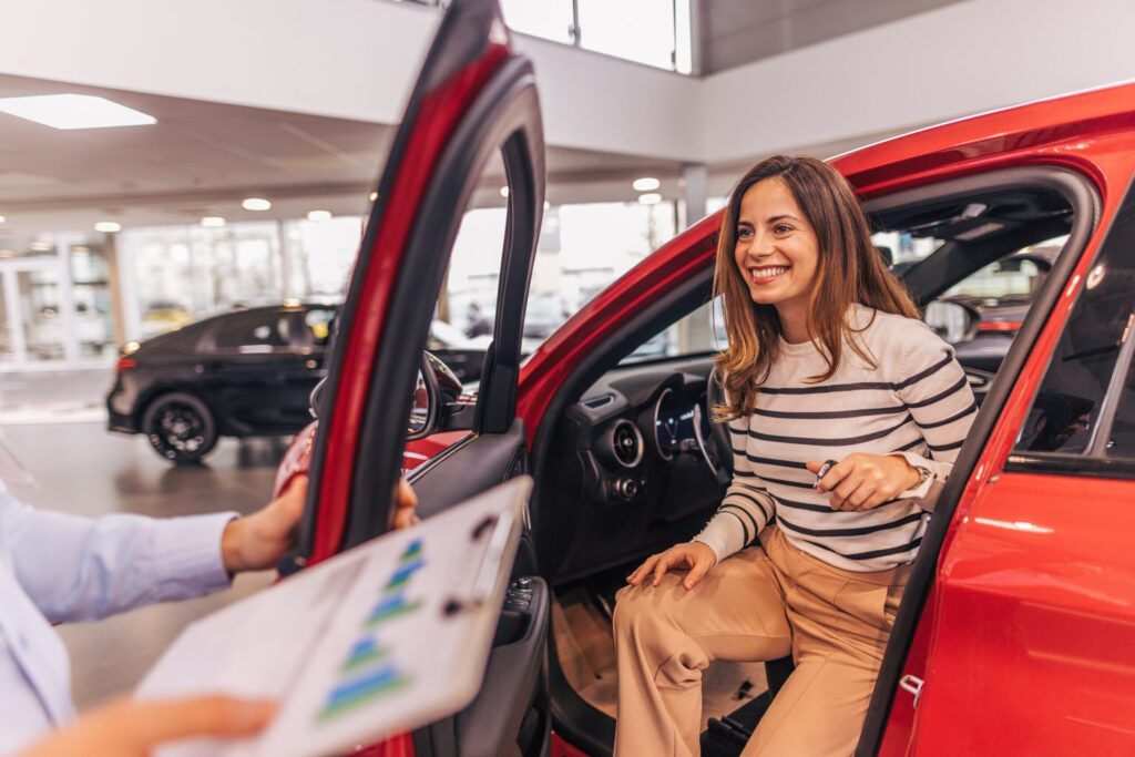 women in drivers seat of new red car in the showroom; salesman holding the door open and holding a tablet with data on it.