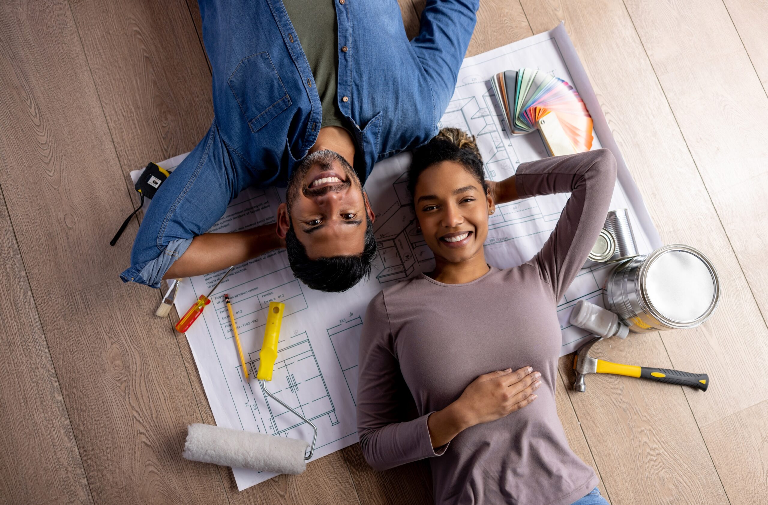 Couple working on a HELOC-funded home renovation project lying down on a wood floor with tools and a blueprint around them