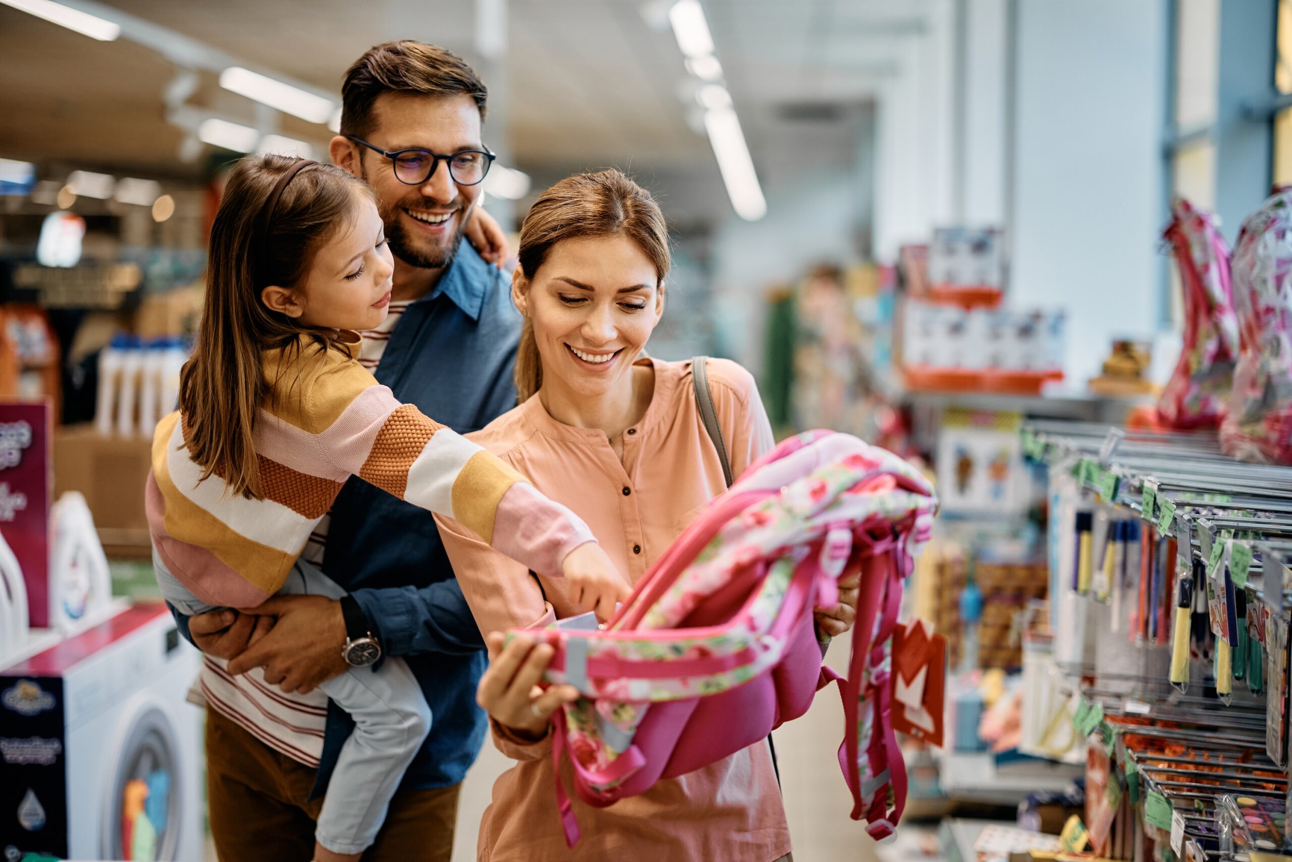 Little girl and her parents navigating back to school expenses and choosing backpack while shopping in a store together.